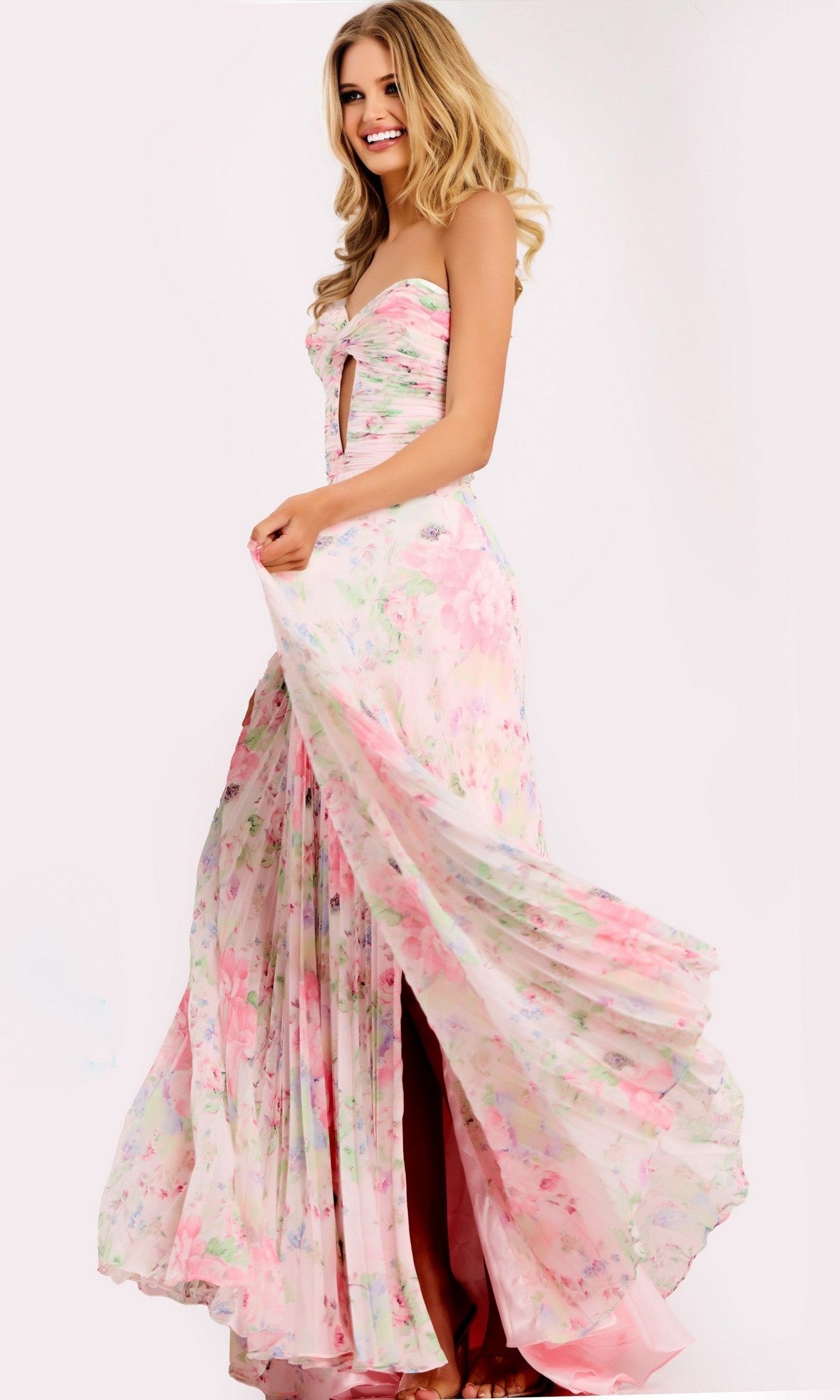 A woman smiles in the Jovani 44518 long prom dress, holding the side of her flowing pink/print chiffon skirt to show its movement—an elegant strapless floral gown perfect for prom.